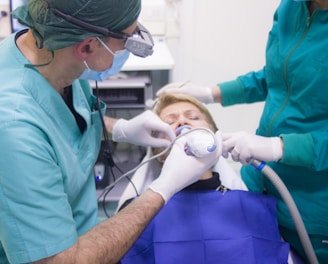 A dental procedure in progress with two medical professionals attending to a patient. The patient is lying back in a dental chair, wearing a blue protective bib. One dentist is using a dental instrument while the other assists, both wearing surgical masks and gloves.
