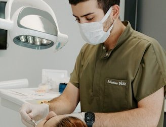 A dentist wearing a mask and green scrubs is attending to a patient lying in a dental chair. The dentist is focused on a procedure, using dental instruments under a bright overhead light. The patient has reddish-brown hair tied back and appears relaxed. Clinical tools and supplies are visible in the background, creating a professional and clean atmosphere.
