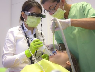 Two dental professionals perform a procedure on a patient. One is wearing specialized eyewear and is focused on using a dental instrument. The patient lies in a dental chair, wearing a protective bib.