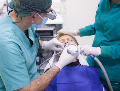 A dental procedure in progress with two medical professionals attending to a patient. The patient is lying back in a dental chair, wearing a blue protective bib. One dentist is using a dental instrument while the other assists, both wearing surgical masks and gloves.