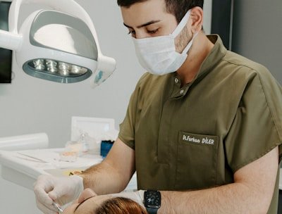 A dentist wearing a mask and green scrubs is attending to a patient lying in a dental chair. The dentist is focused on a procedure, using dental instruments under a bright overhead light. The patient has reddish-brown hair tied back and appears relaxed. Clinical tools and supplies are visible in the background, creating a professional and clean atmosphere.