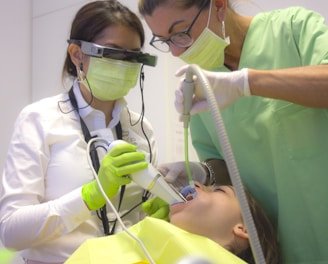 Two dental professionals perform a procedure on a patient. One is wearing specialized eyewear and is focused on using a dental instrument. The patient lies in a dental chair, wearing a protective bib.